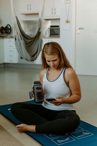 Smiling young female in sportswear holding water bottle and taking selfie on modern mobile phone during home workout