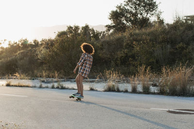 Back view of female hipster with afro hairstyle riding penny board along empty road during sunset