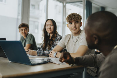 Male teacher discussing with teenage boys and girl sitting at desk in classroom