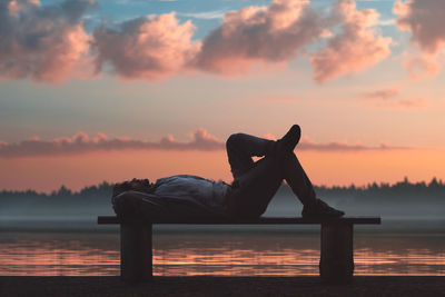 Side view of man lying on bench by lake against sky during sunset