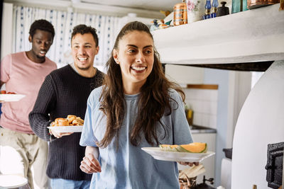 Smiling friends holding food while standing in kitchen at home