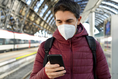 Man wearing mask using phone standing on railroad station