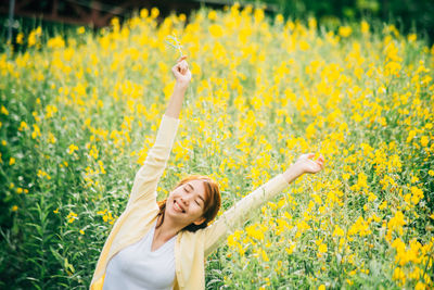 Smiling young woman with arms raised standing against yellow flowering plants