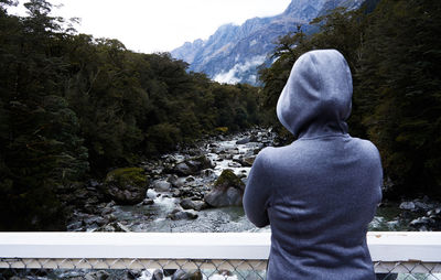 Rear view of person wearing hooded jacket looking at trees and river