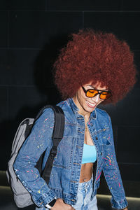 Smiling female with afro hairstyle and trendy outfit and sunglasses looking at the ground while standing on black background with backpack in evening time