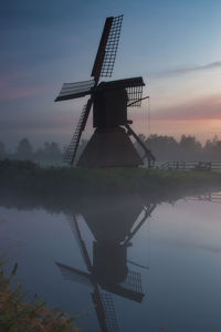 Traditional windmill by lake against sky during sunset