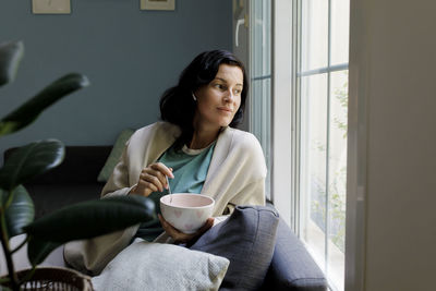 Woman holding bowl looking out through window at home