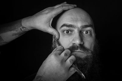 Close-up cropped hands shaving man beard against black background