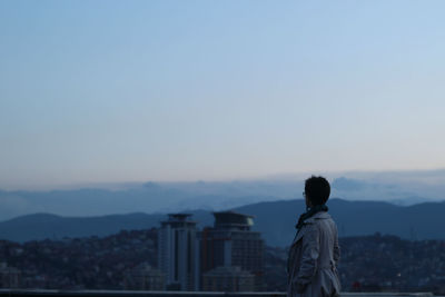 Rear view of man looking at cityscape while standing on rooftop against clear sky