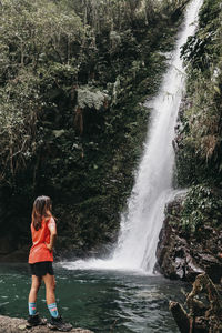 Rear view of woman standing against waterfall in forest