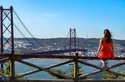 Rear view of woman looking at bridge while sitting on railing against blue sky