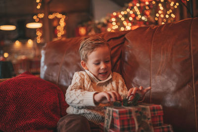 Candid authentic happy child in knitted beige sweater sitting with presents at lodge xmas decorated