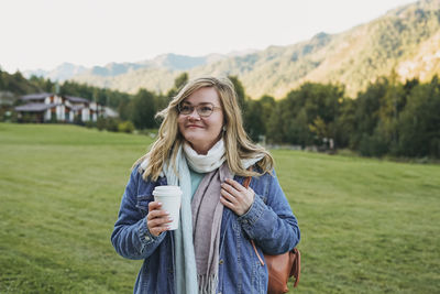 Young woman in jeans coat with paper cup of coffee at chalet cottage village on mountains region