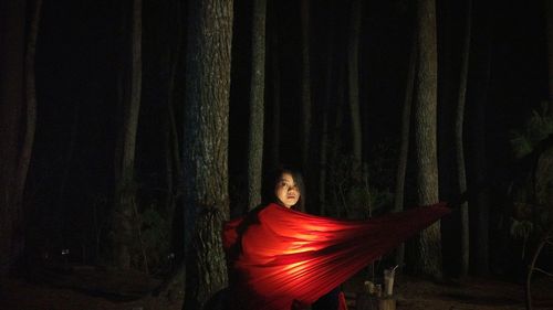 Portrait of young woman sitting in red illuminated hammock at forest during night