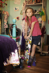 Portrait of confident girl holding skateboard while standing in bedroom