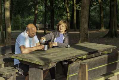 Senior couple sitting in picnic table in forest drkinking water from bottles in metal camping cups