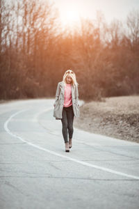 Full length of young woman walking on road against bare trees during sunset