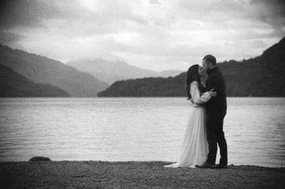 Man embracing woman while standing on lakeshore against sky