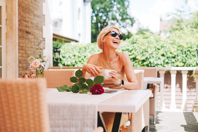 Adult elegant blonde woman in sunglasses beige clothes with cup of coffee and red rose in cafe