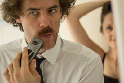 Businessman trimming beard on bathroom while woman looking at him