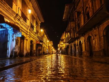 Wet street amidst illuminated buildings in city at night