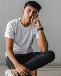 Young man looking away while sitting on seat against wall