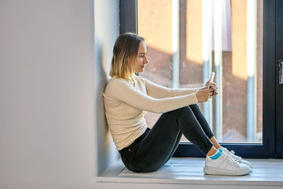 Side view of woman using mobile phone sitting by wall