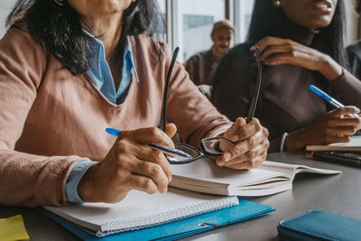 Midsection of mature female student holding eyeglasses while sitting at desk in classroom