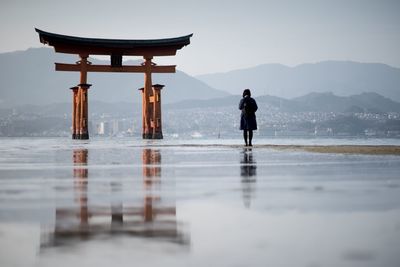 Rear view of woman standing at beach by torii gate against sky