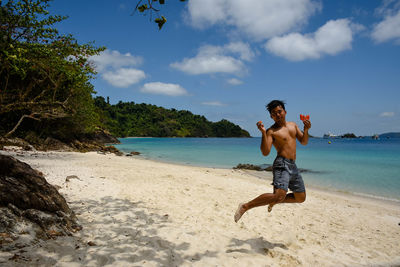 Full length portrait of shirtless man holding fruit while jumping at beach against sky