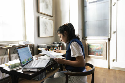 Female designer analyzing documents while sitting on chair in home office