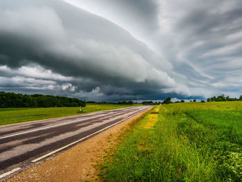 Road passing through field against storm clouds