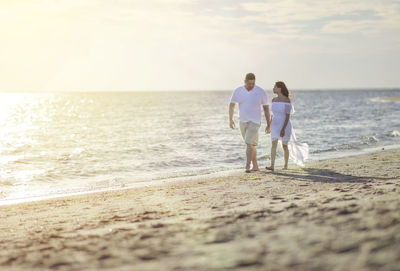 Full length of couple walking on shore at beach against sky during sunset