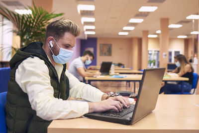 A young boy with a mask sitting and working with his laptop in the library