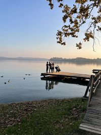 People on pier over lake against sky during sunset