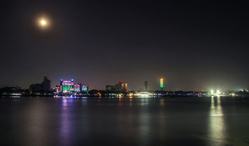 Illuminated buildings by river against sky at night