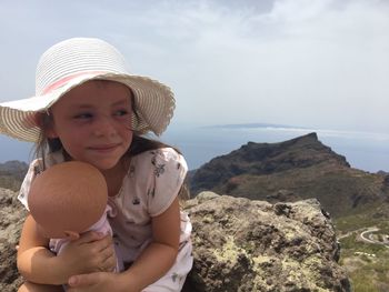 Smiling girl with doll sitting on rock against mountain
