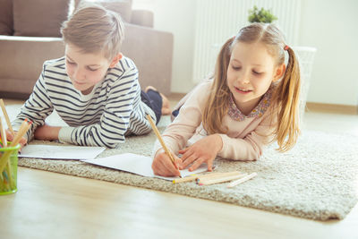 Siblings writing on paper while lying at home