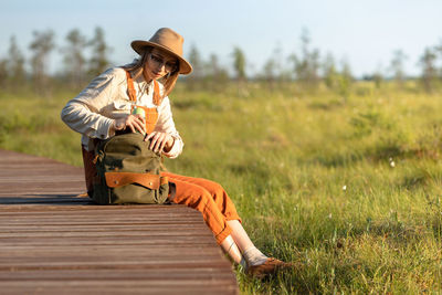 Naturalist woman in hat resting on boardwalk, takes thermos from backpack. ecotourism