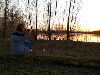 Woman sitting on bare tree against sky during sunset