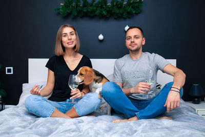 Portrait of couple holding wineglass sitting on bed with dog