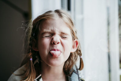 Blond girl sticking tongue out while standing by window at home