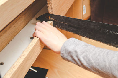 The hand of a young caucasian guy is sawing a board with a saw from the back of a desk in his room.