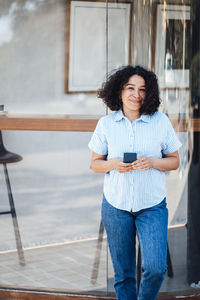 Smiling woman with smart phone leaning on glass wall