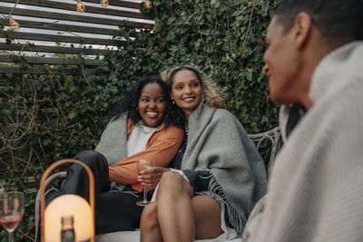 Smiling female friends wrapped in blanket talking with male friend in back yard