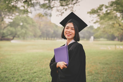 Happy young woman in graduation gown standing on field