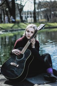 Portrait of young woman with acoustic guitar sitting at lakeshore