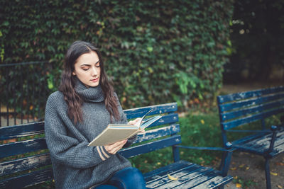 Young woman in a sweater reading a book, sitting on the bench in the park. fall or spring time.