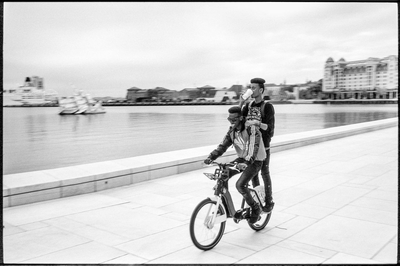 transportation, bicycle, architecture, mode of transportation, real people, water, full length, city, built structure, auto post production filter, people, building exterior, men, day, transfer print, lifestyles, sky, rear view, two people, riding, outdoors