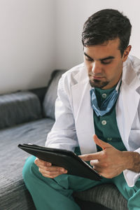 Concentrated male medic in uniform sitting on sofa in clinic and browsing tablet during work